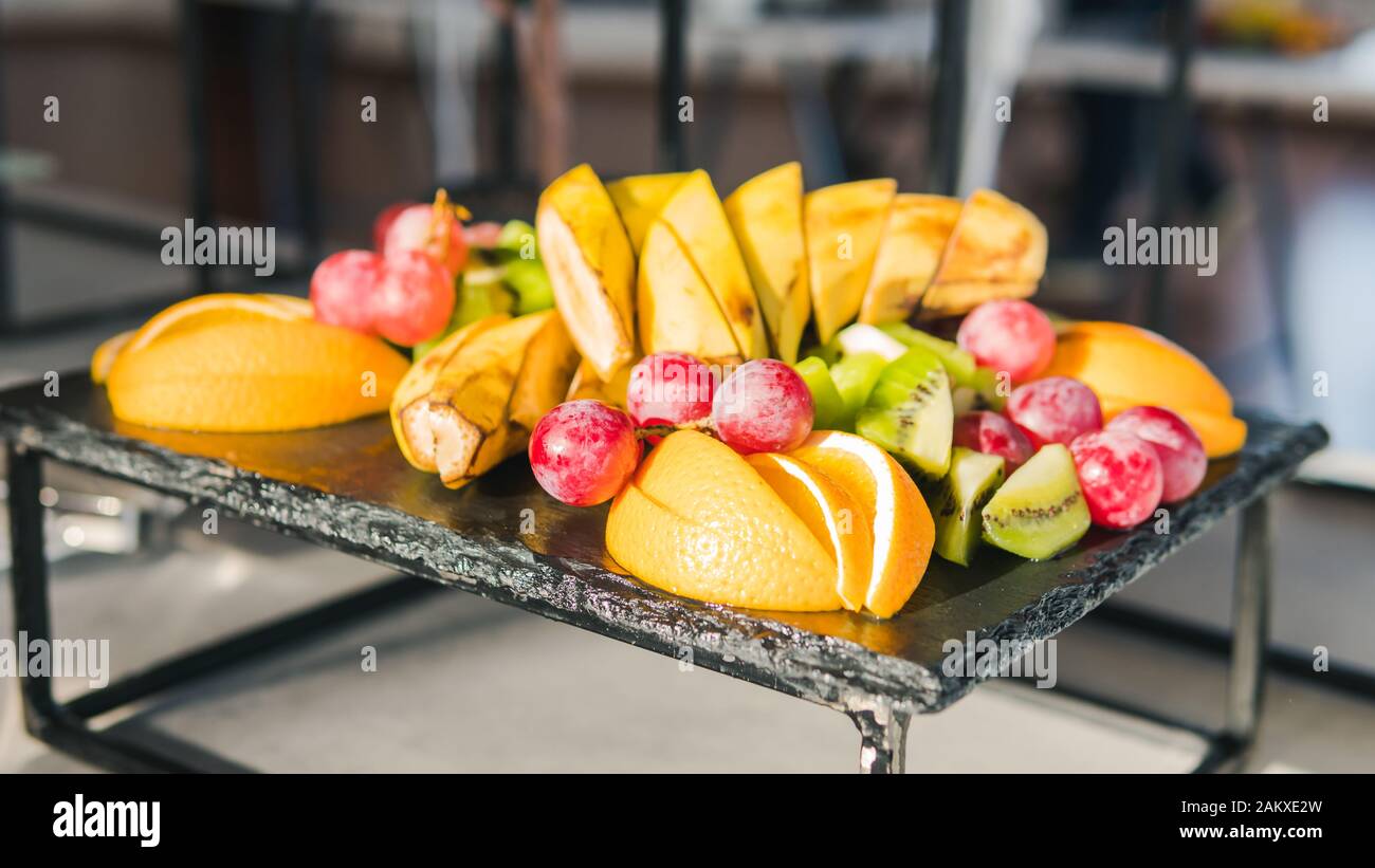 Primo piano di frutta fresca tropicale. Banane, kiwi, arancio e uva sono sul piatto. Concetto di sano servizio di ristorazione e catering Foto Stock