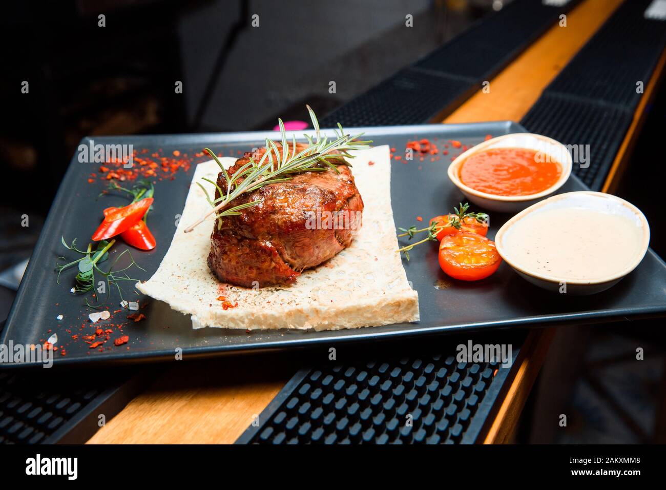 Una bistecca alla griglia succosa con due salse si siede su un piatto nero. Piatti caldi A Base Di Carne, top decorato con un rametto di rosmarino. Foto Stock