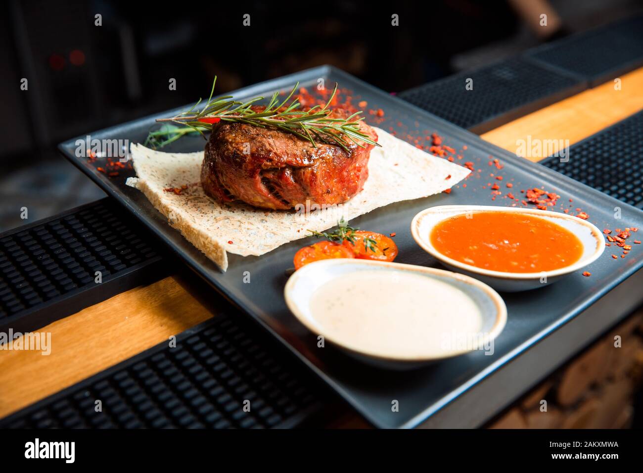 Una bistecca alla griglia succosa con due salse si siede su un piatto nero. Piatti caldi A Base Di Carne, top decorato con un rametto di rosmarino. Foto Stock