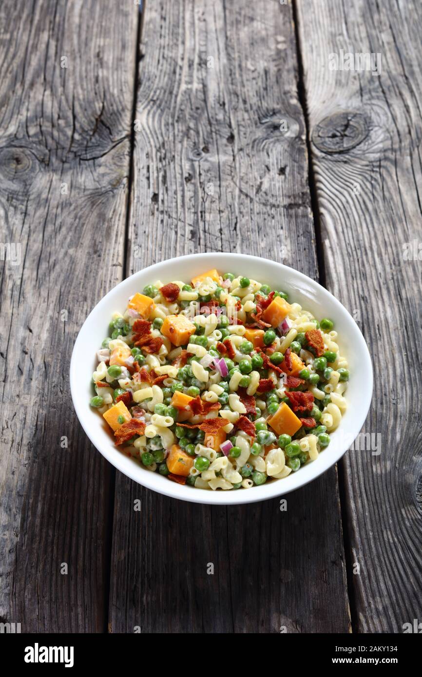 deliziosa insalata di pasta con piselli verdi, pancetta croccante fritta,  formaggio cheddar a cubetti e cipolla rossa in una ciotola bianca su un  tavolo rustico in legno, vista verticale Foto stock 