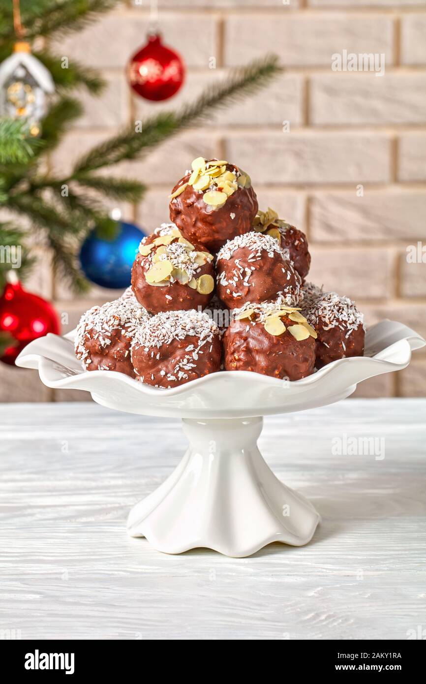 Palline di mandorle di cocco al cioccolato su una base per torte con un albero di Natale e caraffa con caramelle sullo sfondo della parete di mattoni, vista verticale dall'alto, c. Foto Stock