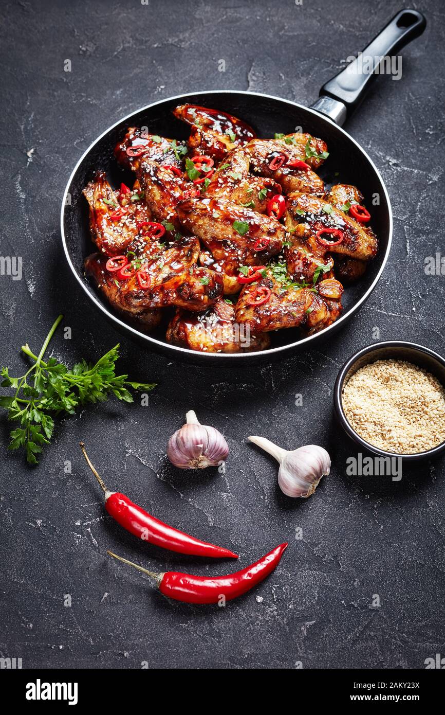 ali di pollo croccanti teriyaki cosparse di semi di sesamo, pezzi di peperoncino e prezzemolo in una padella su un tavolo di cemento, vista verticale dall'alto Foto Stock