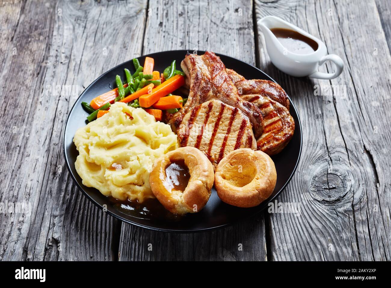 Primo piano di arrosto di domenica inglese con costolette di maiale alla griglia su ossa, purè di patate, fagioli verdi, carote, budini dello Yorkshire e salsa di cipolle su un blac Foto Stock