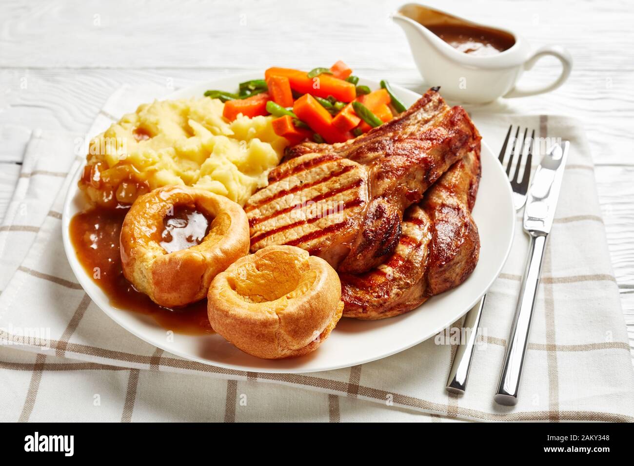 Costolette di maiale alla griglia su ossa, purè di patate, fagioli verdi, carote, budini dello Yorkshire e salsa di cipolle su un piatto bianco su un tavolo di legno Foto Stock