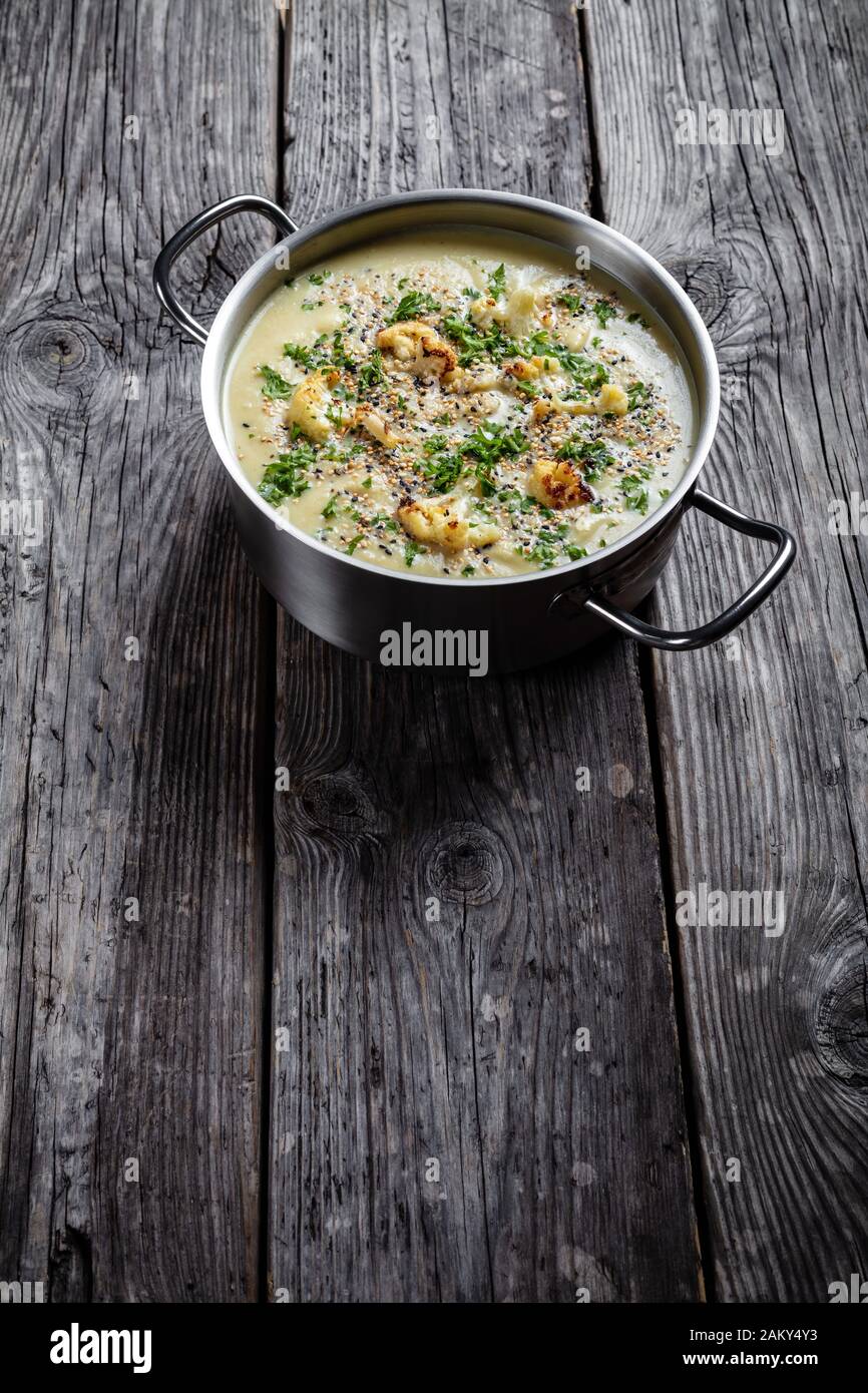 Zuppa cremosa di cavolfiore all'aglio arrosto con formaggio asiago e un croccante di semi di quinoa e chia in un piatto di brodo su un rustico tavolo di legno, v. Foto Stock