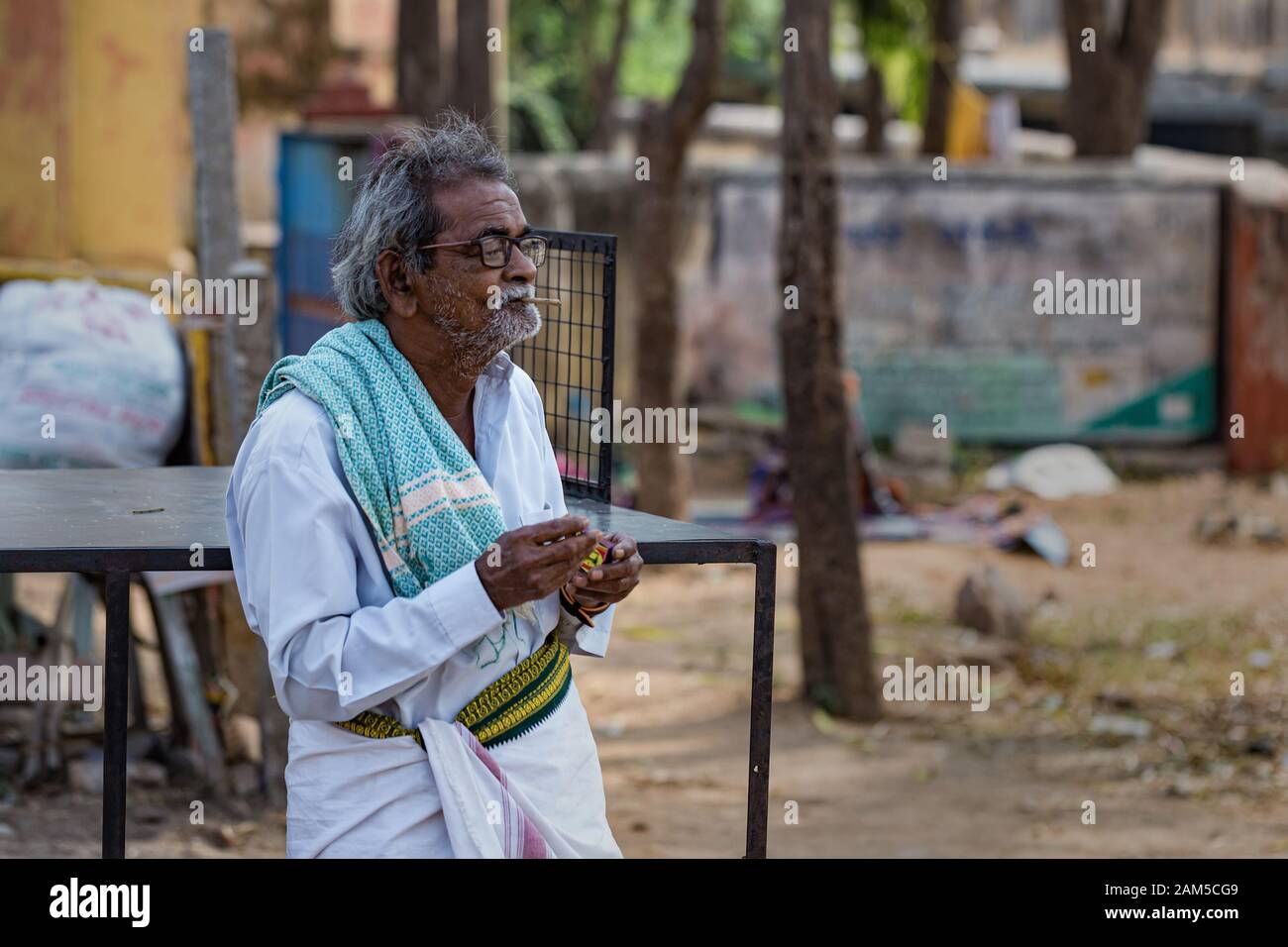Hampi, Karnataka, INDIA - 16 GENNAIO 2018: L'anziano indiano negli occhiali illumina una sigaretta. Residente in india. Un uomo in una camicia bianca e dhoti Foto Stock