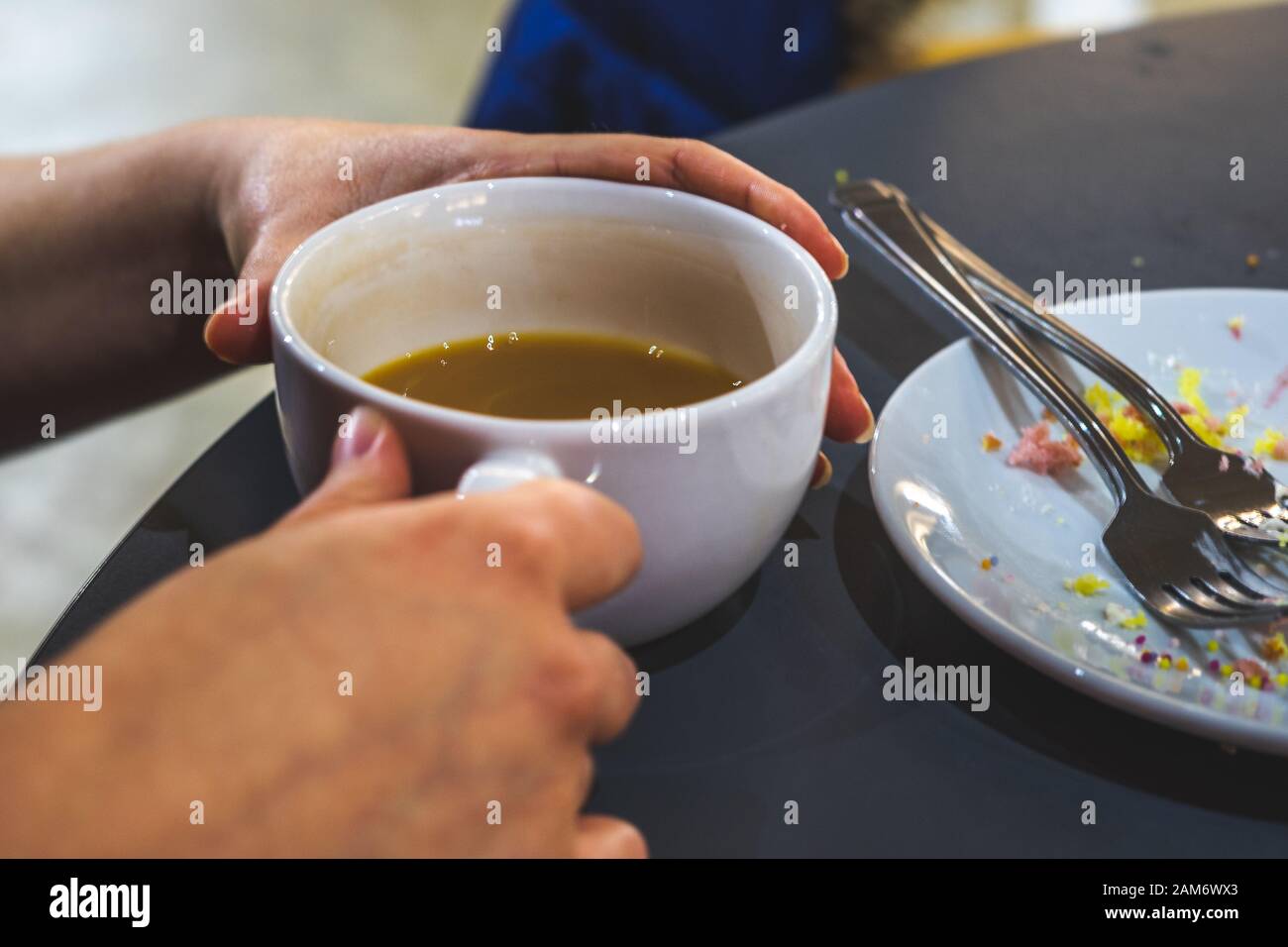 mani che tengono una tazza mezza bevuta di tè con il piatto vicino vicino con le briciole della torta sopra Foto Stock