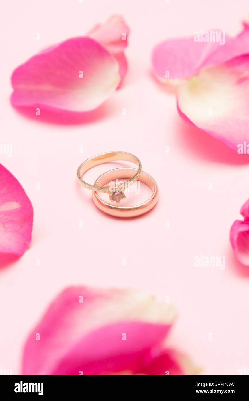 Anello di nozze e anello di fidanzamento d'oro con gemstone intorno ai petali di rose sullo sfondo rosa pastello. Concetto di matrimonio. Buon giorno di San Valentino. Foto Stock