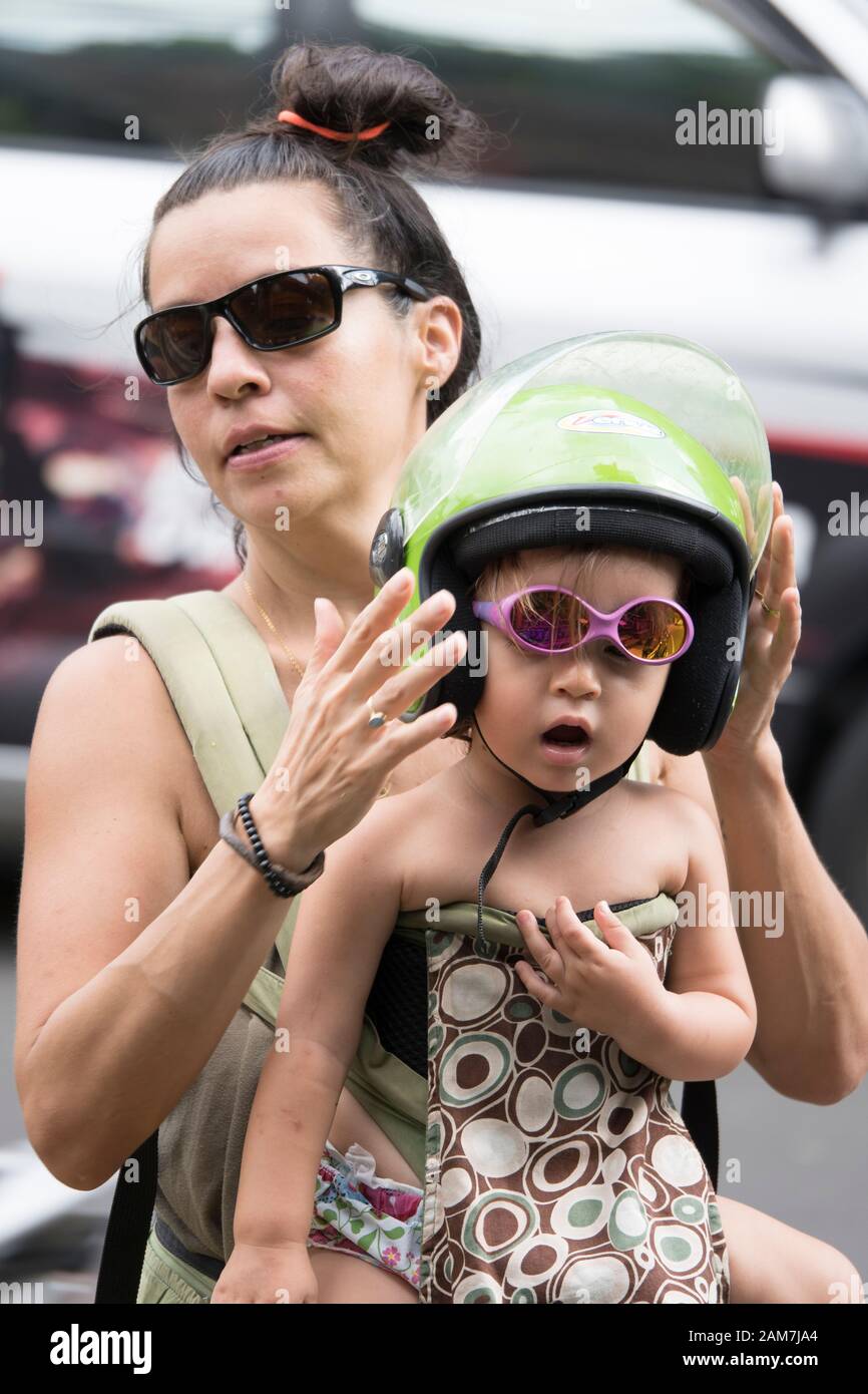 Giovane madre turistica che mette un casco verde di scooter di calce sul suo vetro di sole rosa che indossa cute, figlia del toddler prima di ottenere sullo scooter. Foto Stock