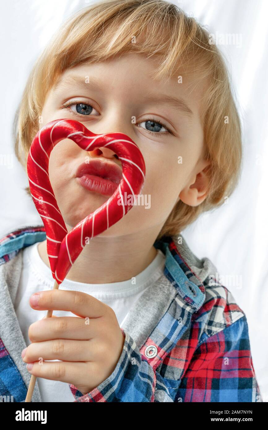 Carino ragazzino con dolcetti rossi a forma di cuore, sfondo bianco. Un bellissimo bambino mangia dolci. San Valentino, amore concetto. Foto Stock