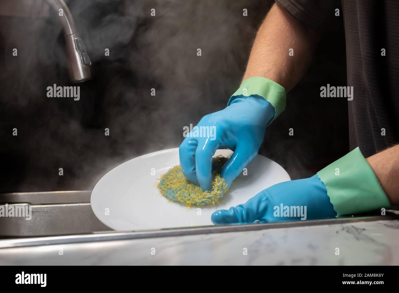 Primo piano dei piatti per il lavaggio delle mani di un modello in acqua calda fumante con guanti in gomma blu chiaro con polsini anti-gocciolamento. Foto Stock