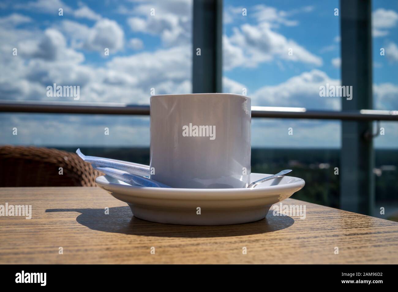 Una tazza bianca di tè o caffè con un cucchiaio e zucchero sul piattino di ceramica, servita nel ristorante. Vista laterale ravvicinata sul tavolo in legno contro la finestra luminosa Foto Stock
