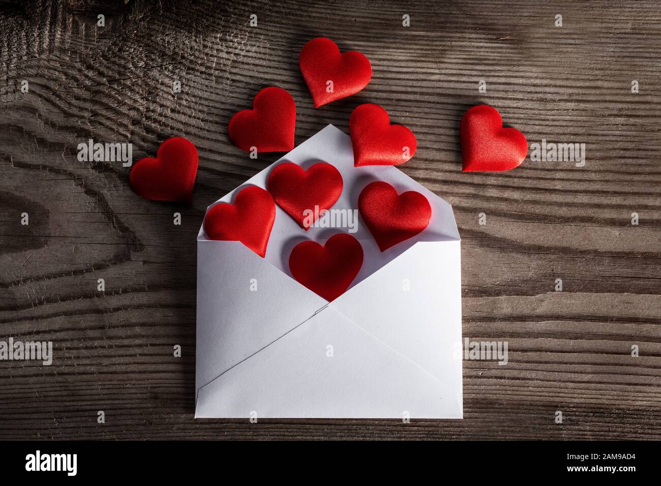 Lettera d'amore di giorno di San Valentino, busta con heap rosso dei cuori sparsi su sfondo di legno. Foto Stock