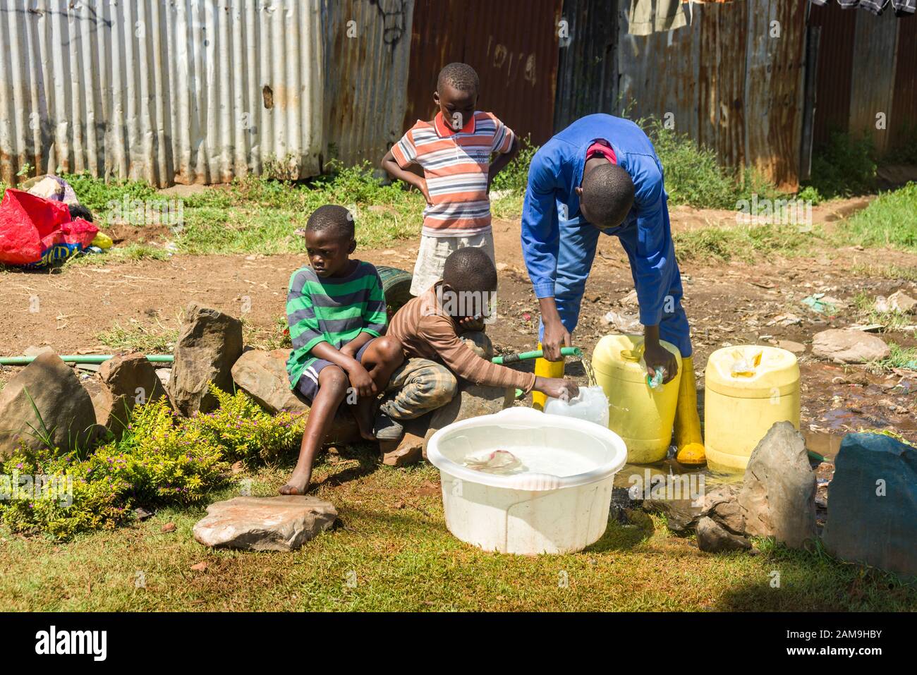 Un giovane uomo e alcuni ragazzi locali riempiono i contenitori d'acqua da un tubo d'acqua dolce, Korogocho slum, Nairobi, Kenya Foto Stock