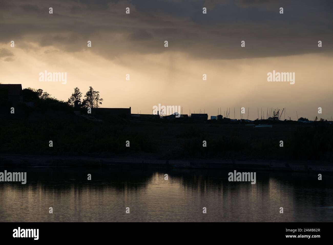 Silhouette di Brancaster Staithes nel nord del Norfolk con nuvole di pioggia che si avvicinano alla luce del sole di prima sera e si riflettono nell'acqua Foto Stock