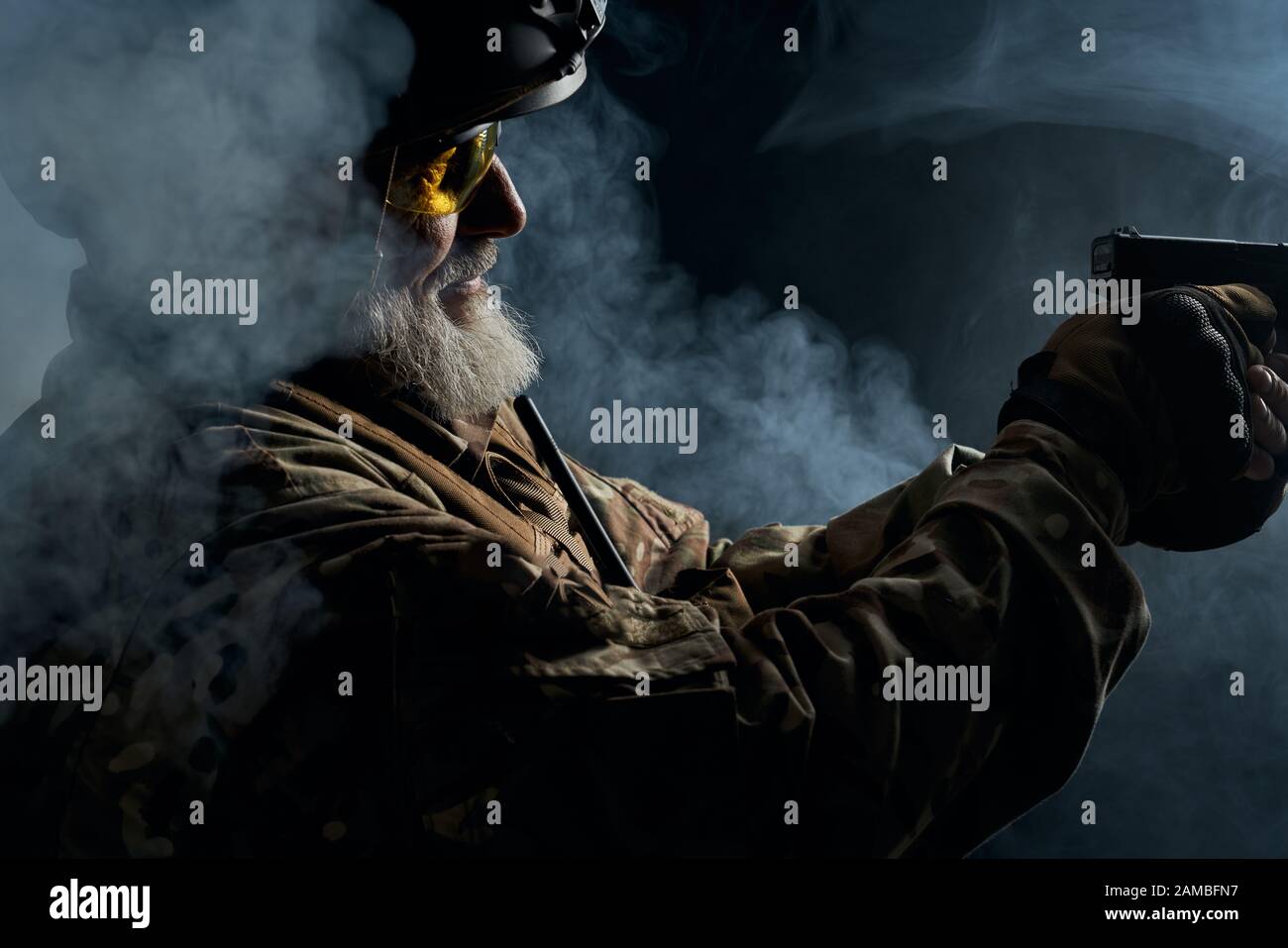 Primo piano di vecchio ufficiale militare che detiene una piccola pistola e mira. Ritratto di veterano bearded in uniforme camouflage e scatto del casco in atmosfera scura fumosa. Concetto di militare. Foto Stock