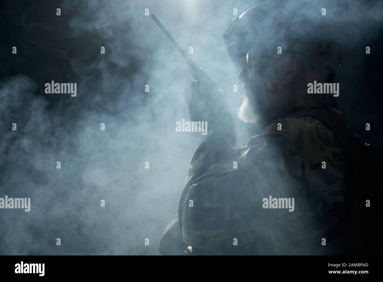 Vista laterale del vecchio soldato militare in uniforme che tiene la stazione radio portatile in fumo. Primo piano del comandante maschile in uniforme camouflage dando ordini in atmosfera scura. Concetto di esercito, militare. Foto Stock