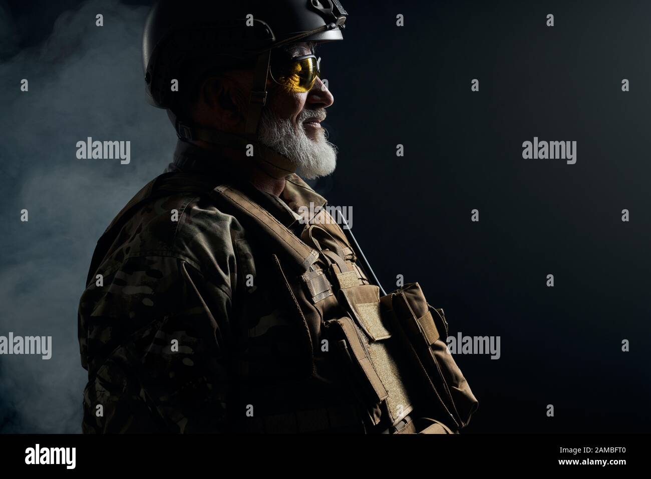 Vista laterale del vecchio ufficiale militare che guarda lontano. Crop ritratto di veterano bearded in uniforme camouflage, occhiali e casco che posano in atmosfera scura fumosa. Concetto di militare, esercito. Foto Stock