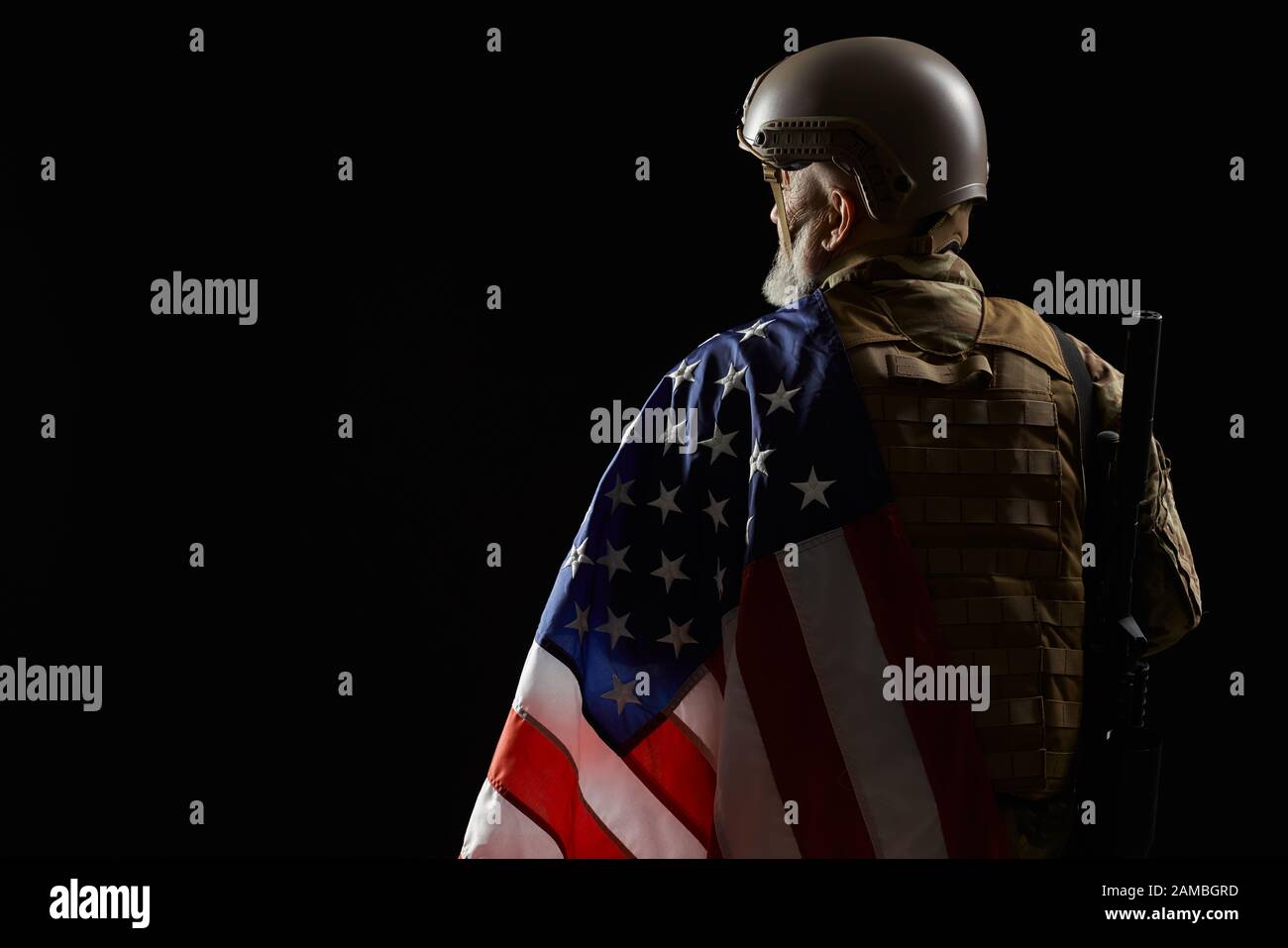 Vista posteriore del veterano militare americano con bandiera e fucile sulla spalla. Ritratto di vecchio ufficiale maschio in incognito in uniforme camuffata che posa in un'atmosfera scura. Concetto di militare, esercito. Foto Stock