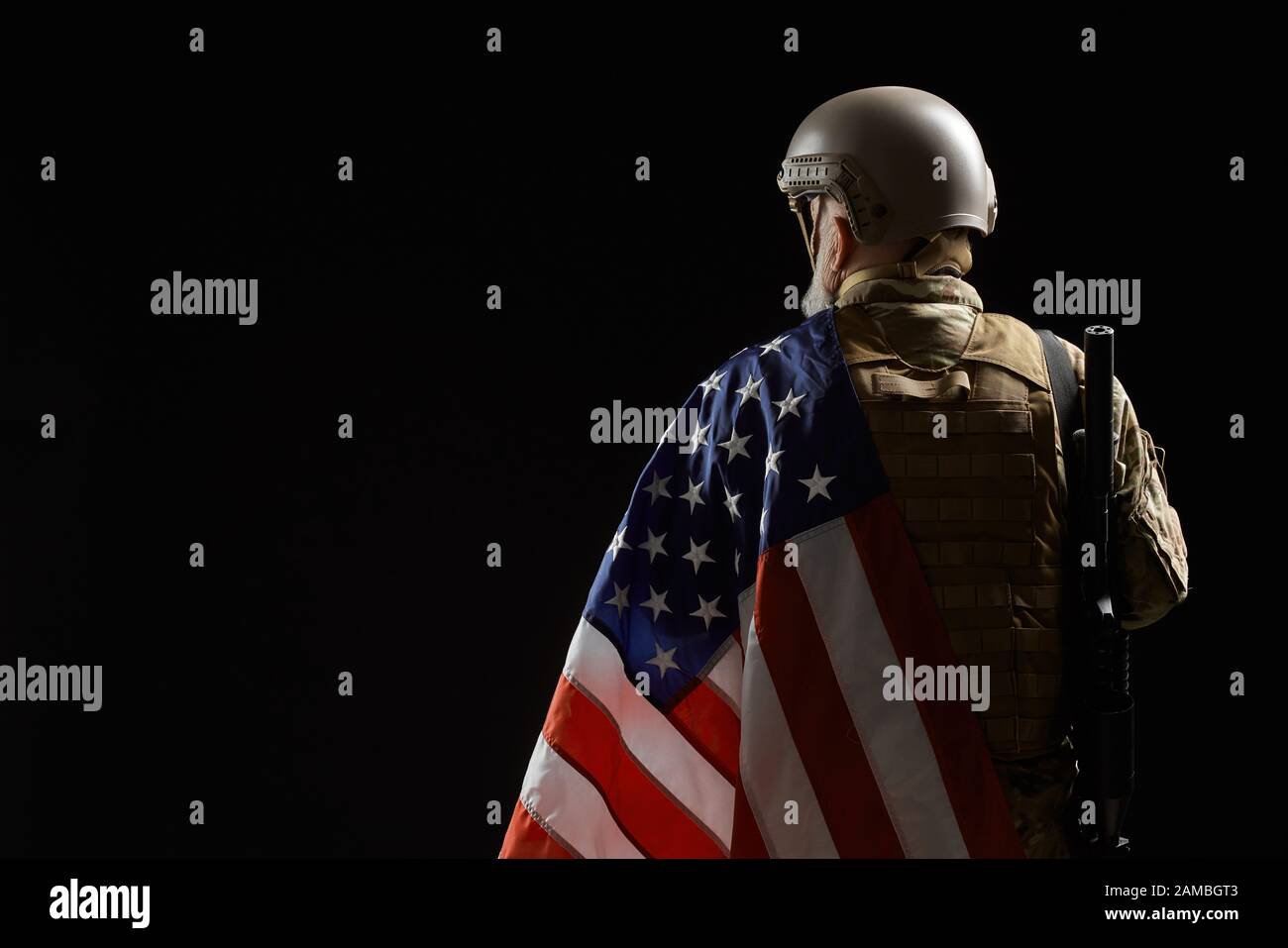 Vista posteriore del veterano militare americano con bandiera e fucile sulla spalla. Ritratto di vecchio ufficiale maschile in incognito in uniforme posa in un'atmosfera buia. Concetto di militare, esercito. Foto Stock