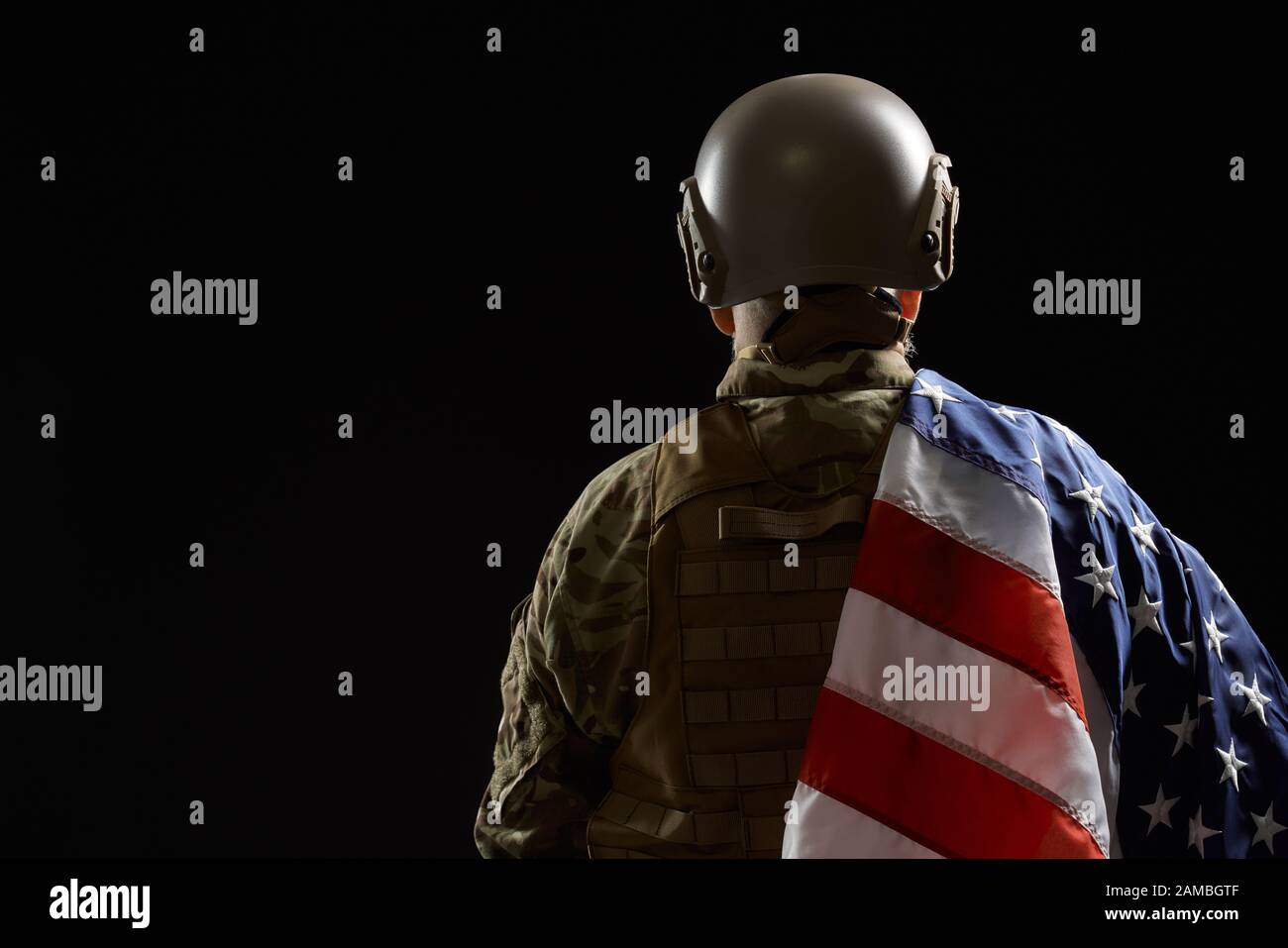 Vista posteriore del veterano militare americano che tiene la bandiera sulla spalla. Ritratto di vecchio ufficiale maschile in incognito in uniforme camouflage posa in un'atmosfera buia. Concetto di militare, esercito. Foto Stock