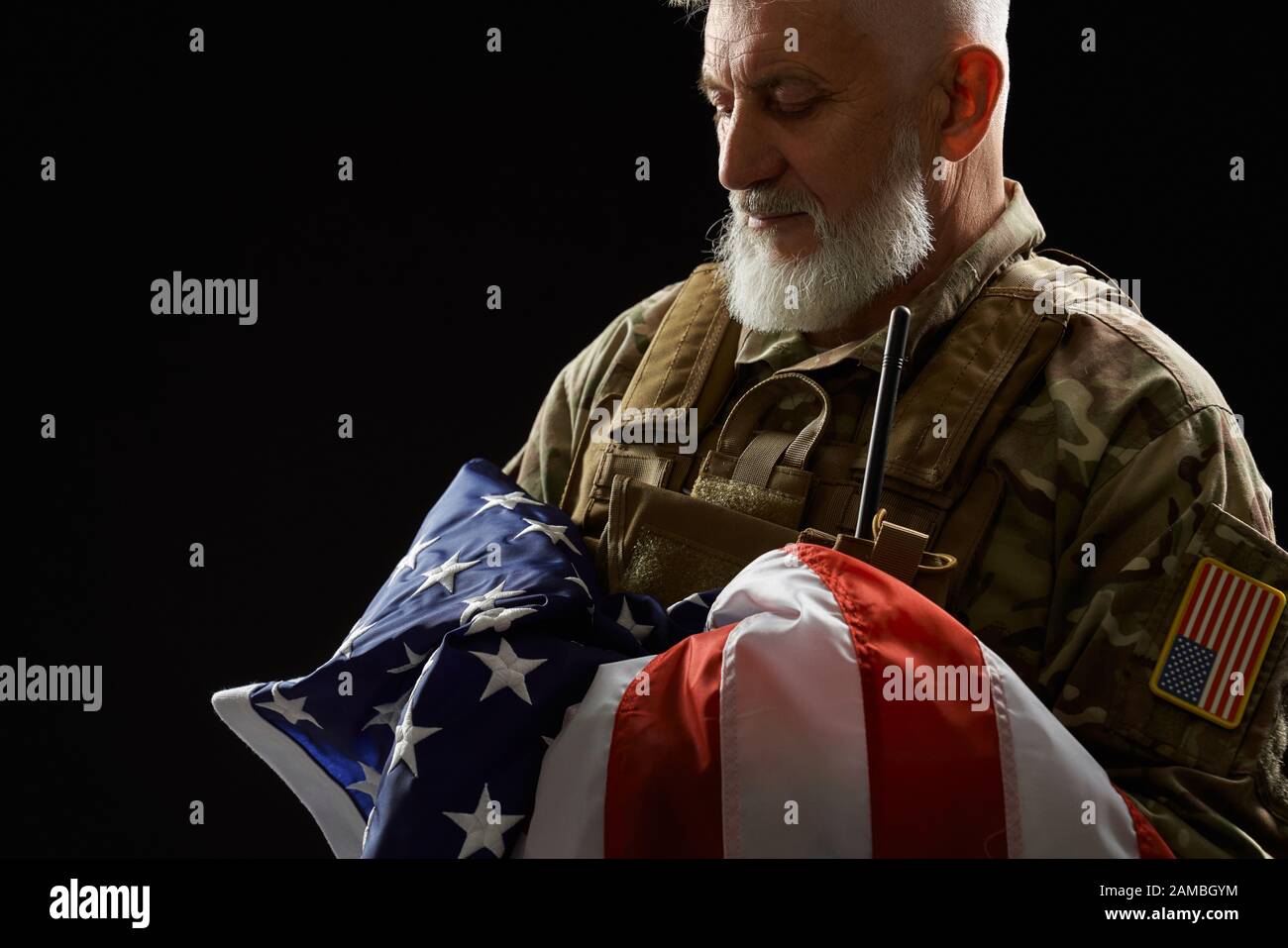Raccolto di bearded veterano militare americano che tiene la bandiera in armi. Ritratto di vecchio ufficiale maschile orgoglioso in uniforme camouflage posa in un'atmosfera buia. Concetto di militare, patriottismo. Foto Stock
