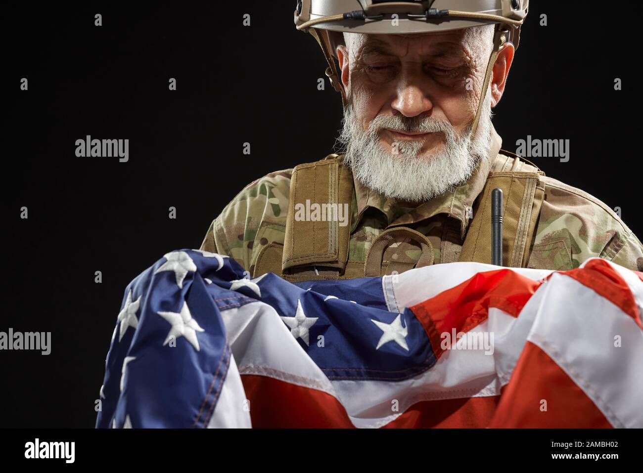 Raccolto di bearded veterano militare americano che tiene la bandiera in armi. Ritratto di vecchio ufficiale maschile orgoglioso in uniforme camuffata e casco in posa in un'atmosfera buia. Concetto di militare, patriottismo. Foto Stock