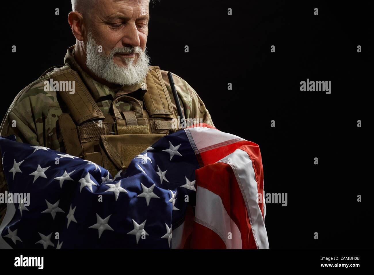 Raccolto di bearded veterano militare americano che tiene la bandiera in armi. Ritratto di vecchio ufficiale maschile orgoglioso in uniforme camouflage posa in un'atmosfera buia. Concetto di militare, patriottismo. Foto Stock