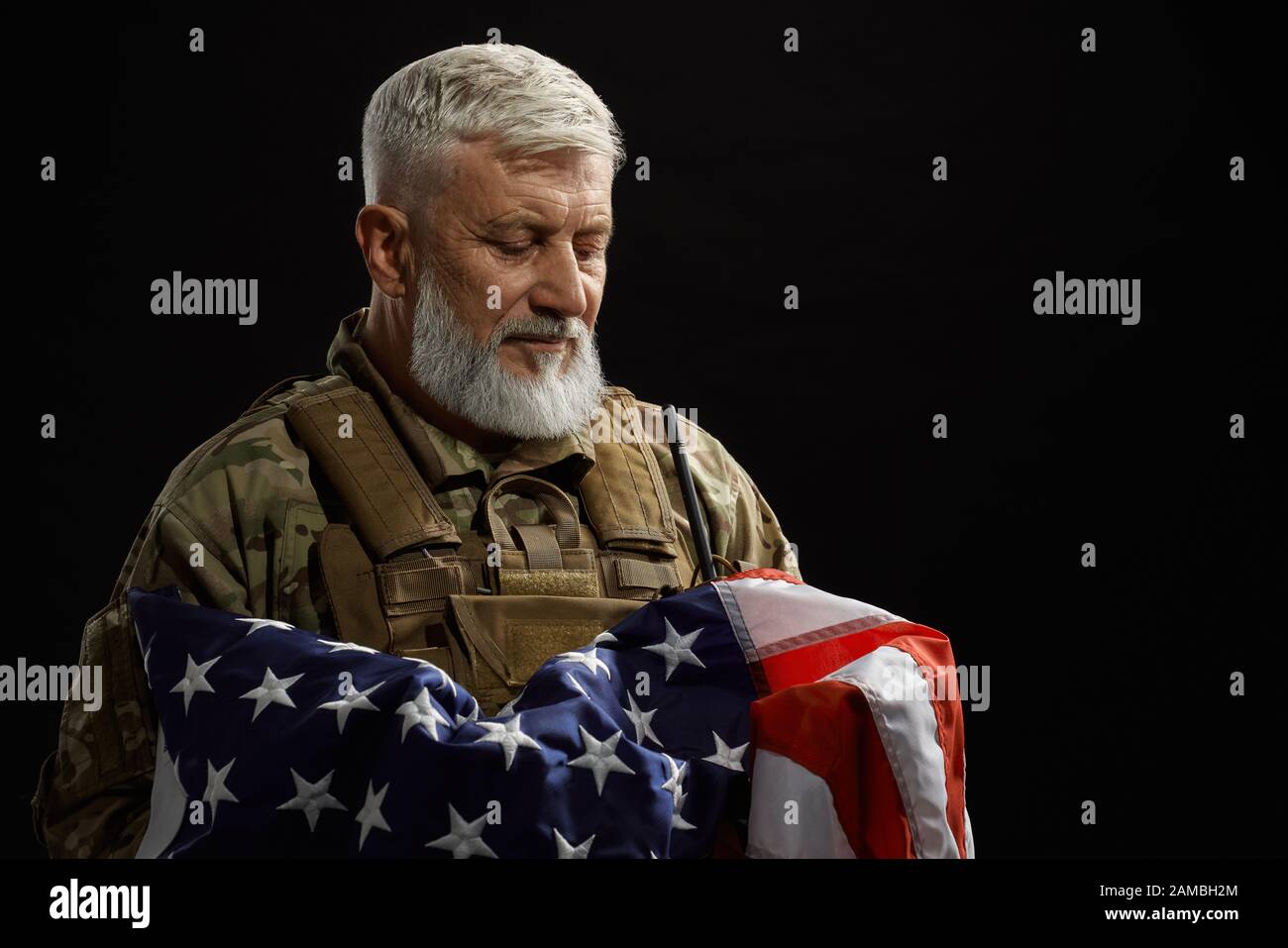 Vista frontale del veterano militare americano con bandiera a braccio. Ritratto di vecchio ufficiale maschile orgoglioso in uniforme camouflage posa in un'atmosfera buia. Concetto di militare, patriottismo. Foto Stock