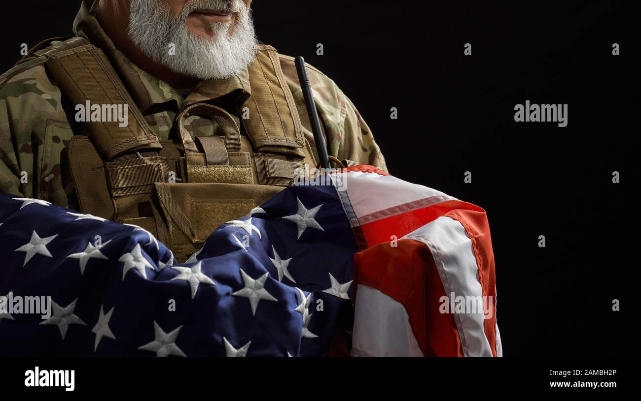 Primo piano di un veterano militare americano con bandiera a braccio. Ritratto di incognito vecchio fiero ufficiale maschile in uniforme camouflage posa in un'atmosfera scura. Concetto di militare, patriottismo. Foto Stock