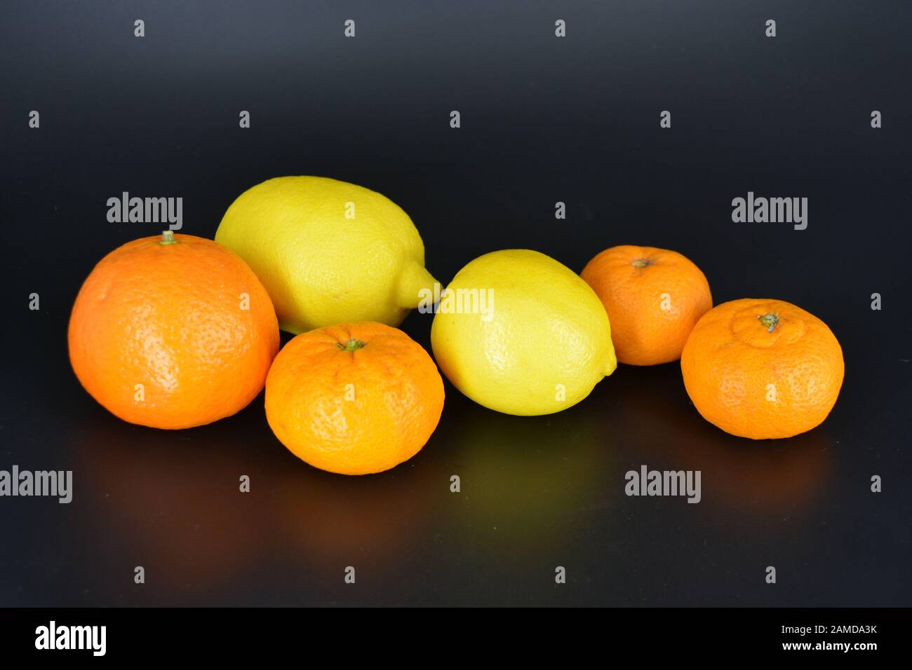 Frutta fresca e brillante, due limoni gialli, tre piccoli mandarini arancioni, una grande mineralola si trova su uno sfondo di plastica nera. Delizioso e dolce. Foto Stock