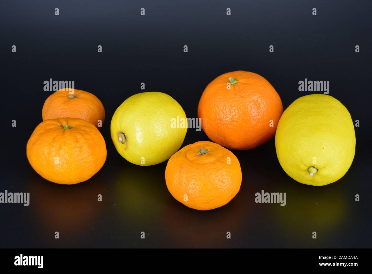 Frutta fresca e brillante, due limoni gialli, tre piccoli mandarini arancioni, una grande mineralola si trova su uno sfondo di plastica nera. Delizioso e dolce. Foto Stock
