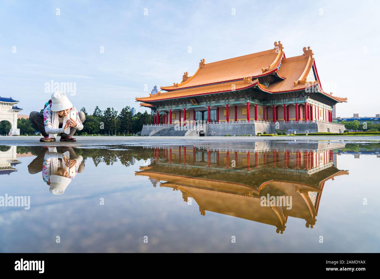 Il turista che prende la foto di basso angolo del riflesso della Sala commemorativa di Chiang Kai-Shek in una pozza di acqua. Edificio del Teatro Nazionale sullo sfondo Foto Stock