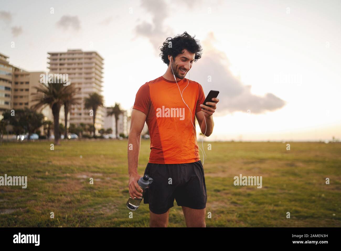 Allegro ritratto di un giovane sportivo che guarda lo schermo del suo smartphone e ascolta la musica negli auricolari bianchi mentre si trova in posizione di parco Foto Stock