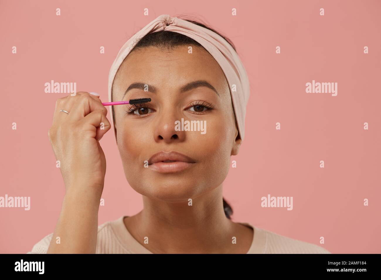 Testa e spalle ritratto di bella razza mista donna applicazione mascara mentre mettere sul trucco al mattino in piedi contro polvere rosa sfondo, copia spazio Foto Stock