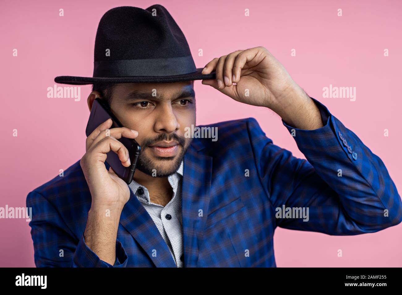 Ritratto di closeup dell'imprenditore indiano elegante che indossa l'abito blu controllato, cappello toccante, che parla sul telefono mobile con espressione seria mentre si siede Foto Stock
