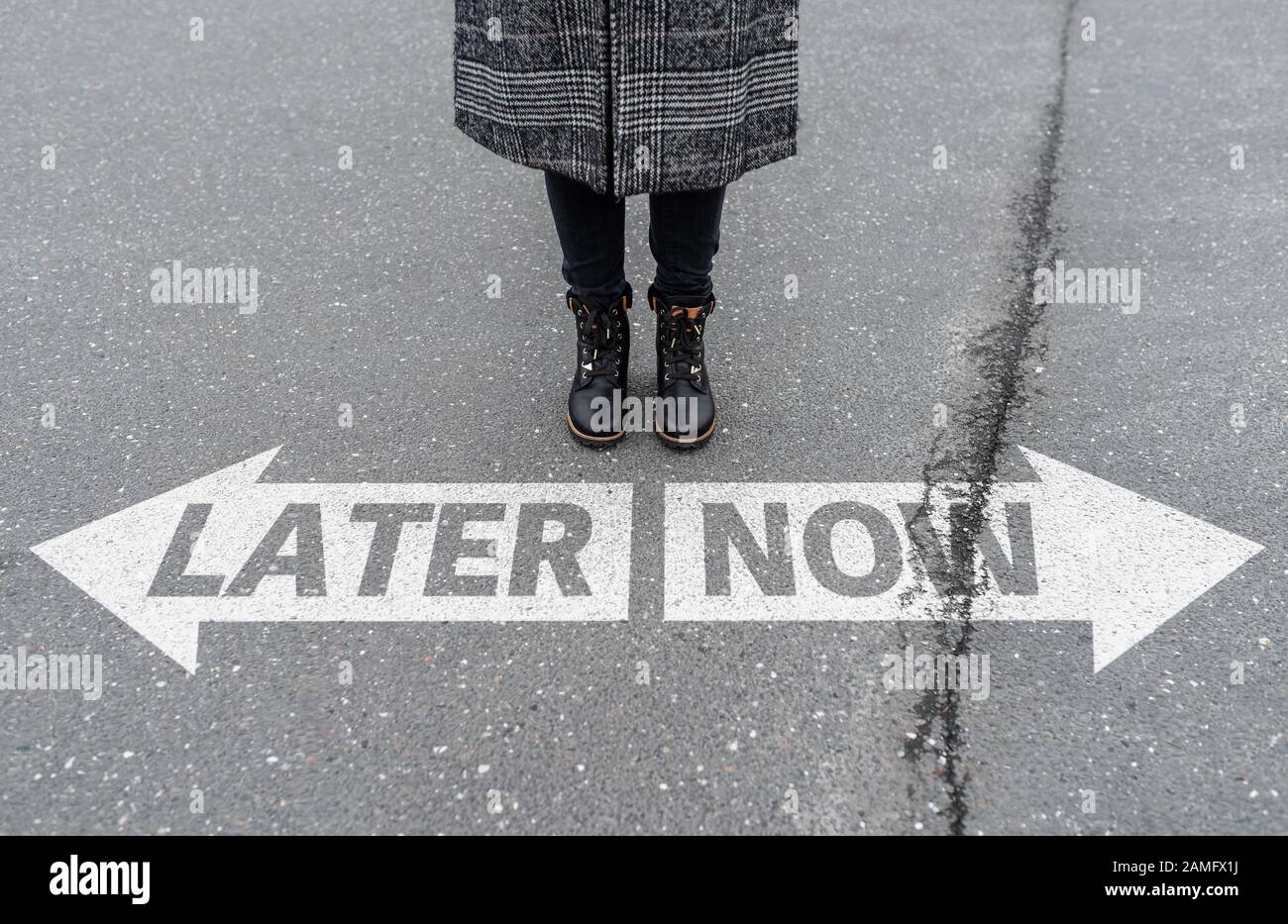 Gambe di donna in piedi dietro la freccia strada marcatura con il testo ORA e PIÙ TARDI, chiamata ad azione e procrastination concetto Foto Stock