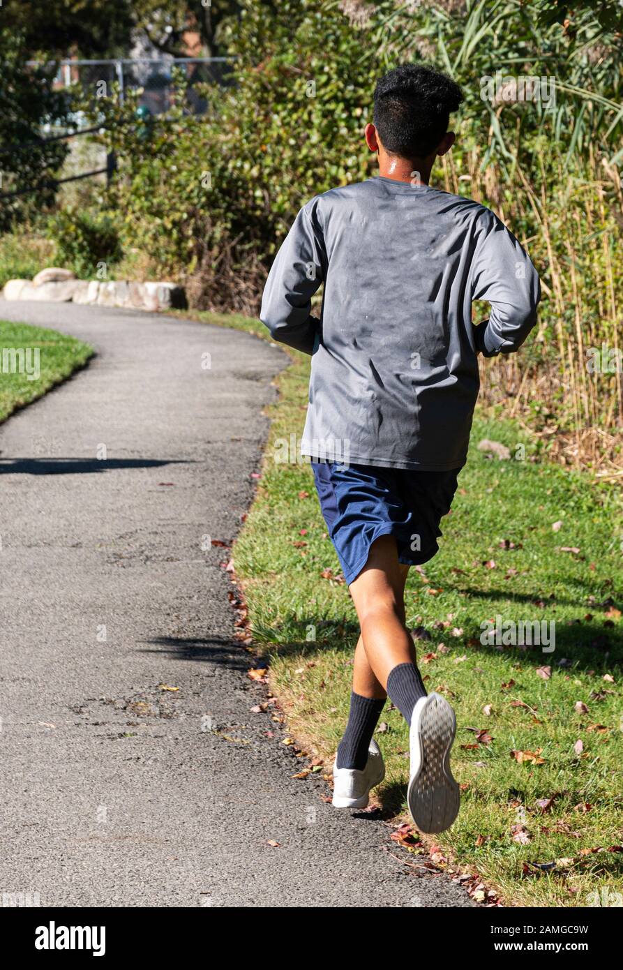 Vista posteriore di un corridore di fondo di scuola superiore che corre su un percorso catrame in un parco con alberi e cespugli sulla sua destra, Foto Stock