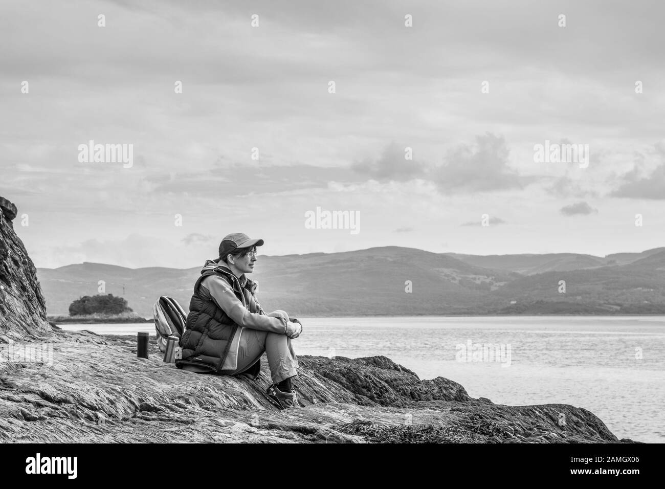 Vista laterale monocromatica di una donna britannica isolata in berretto sportivo e attrezzatura da passeggio, seduta su rocce costiere sulla spiaggia, guardando verso il mare, nel profondo del pensiero. Isolamento. Foto Stock
