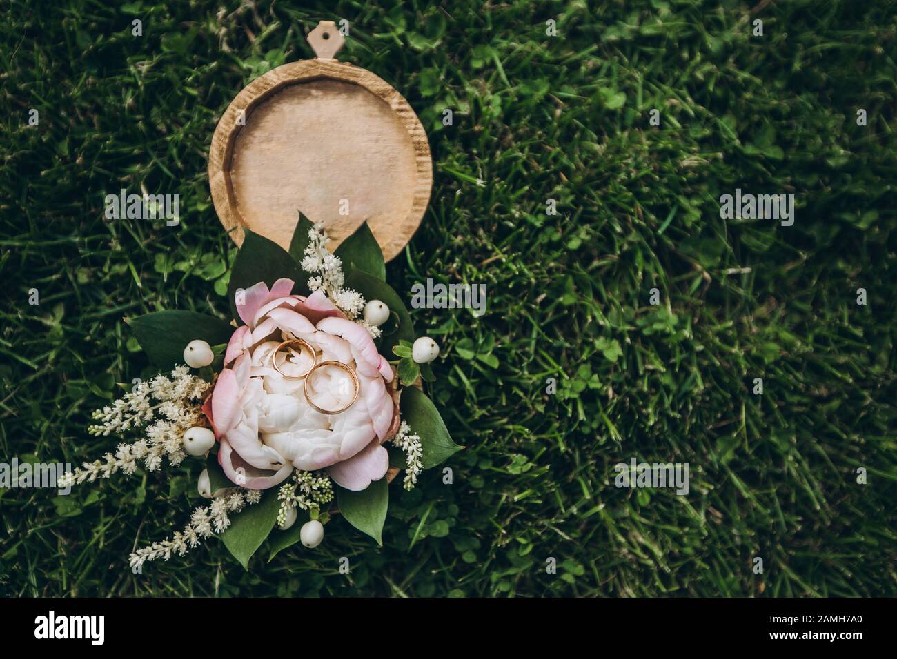 Scatola rotonda in legno con fiori tenera, verde, rose e anelli da sposa dorati, adagiata su una fresca erba verde. Foto Stock