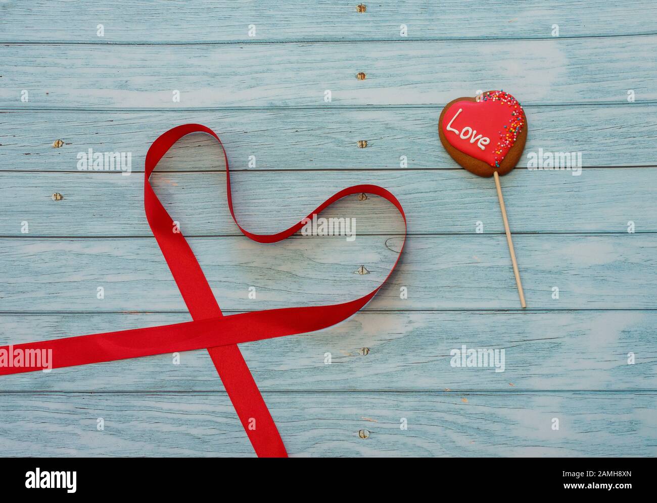 Biscotto decorato a forma di cuore su un bastone con nastro rosso in forma di cuore su sfondo di legno blu. Spazio per il testo. Concept San Valentino. Amore Foto Stock