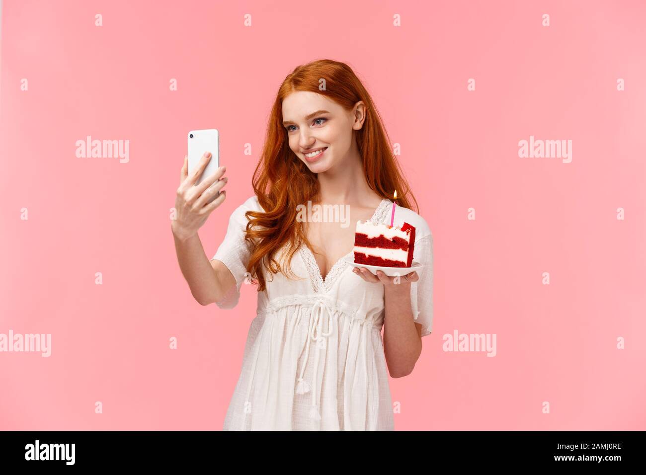 Allurante ragazza caucasica compleanno con capelli lunghi rossi in abito bianco, prendendo selfie per pubblicare i social media internet durante b-day party, tenendo smartphone Foto Stock