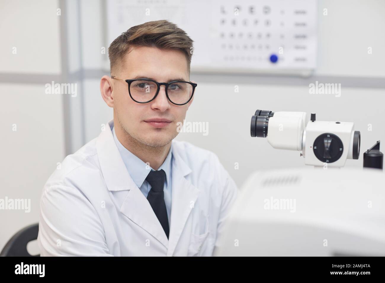 Testa e spalle ritratto di maschio optometrist sorridente alla macchina fotografica mentre posando sul posto di lavoro da apparecchiatura ottica, copia spazio Foto Stock