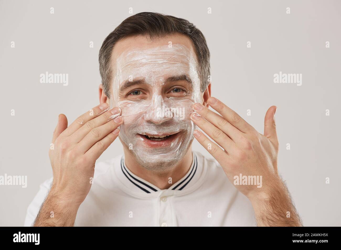 Testa e spalle ritratto di uomo adulto sorridente applicando maschera facciale mentre godendo routine mattutina in piedi su sfondo bianco Foto Stock