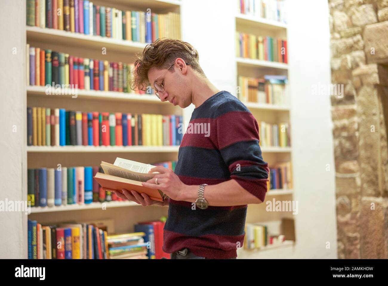 Giovane studente con un orologio e occhiali, leggendo un libro in biblioteca accanto a una libreria. Foto Stock