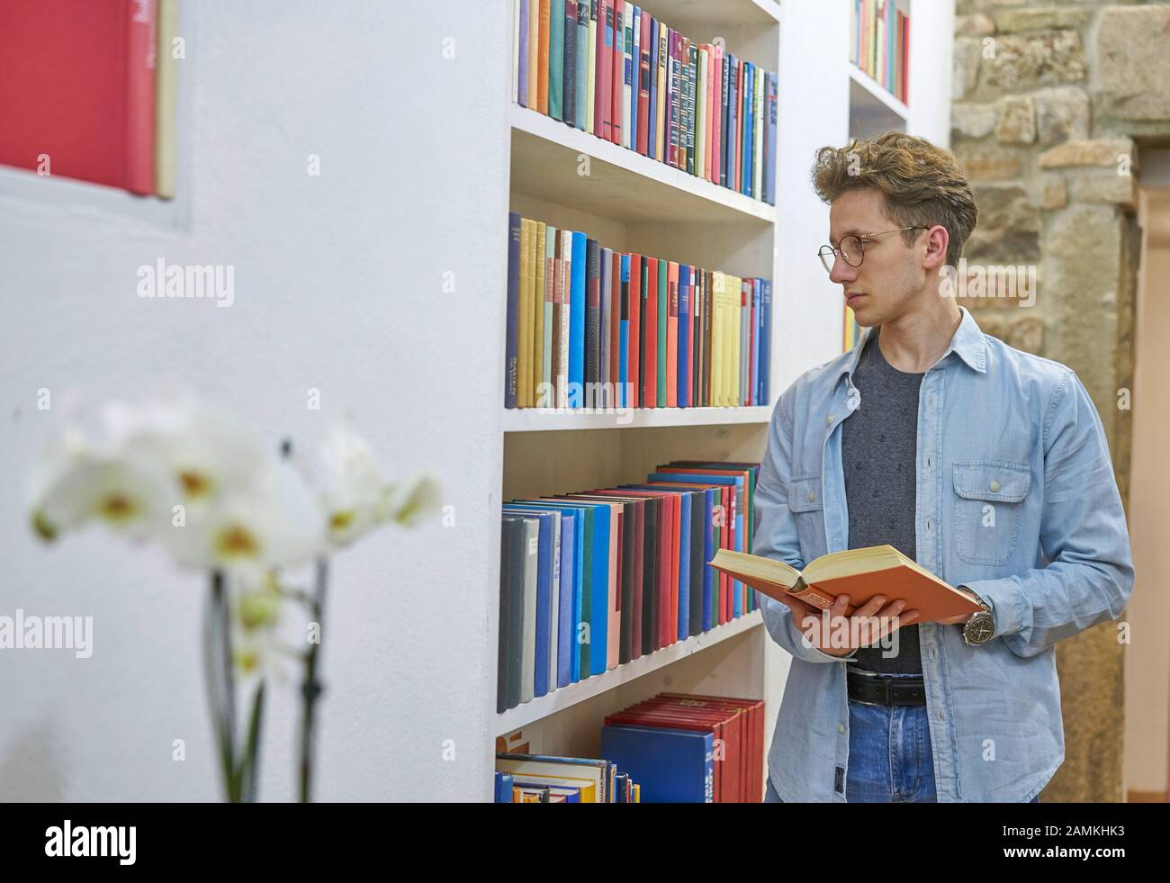 Giovane studente con libro in mano guardando libreria in casa biblioteca. Muro di pietra sullo sfondo. Orchidea in primo piano. Foto Stock