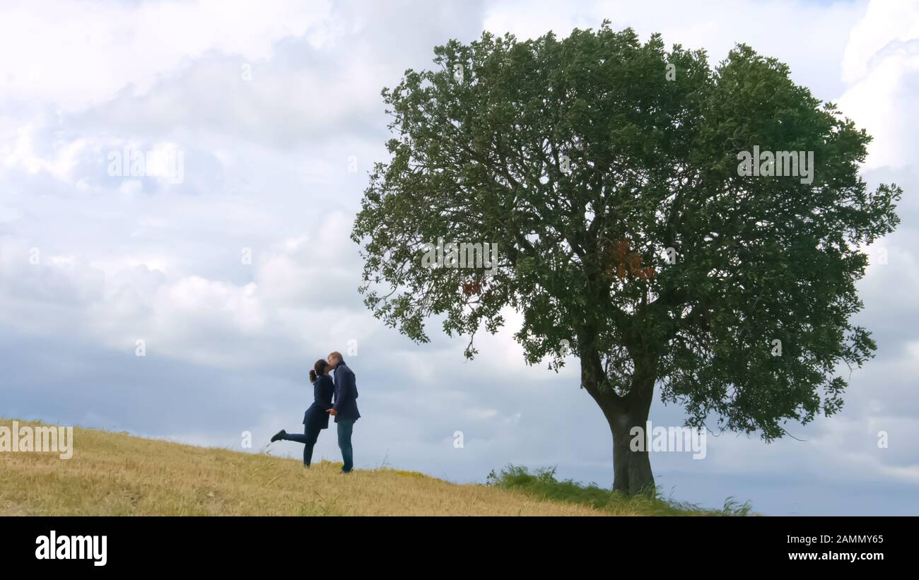 Giovane coppia che abbraccia e bacia sotto il grande albero verde, amore eterno per sempre Foto Stock
