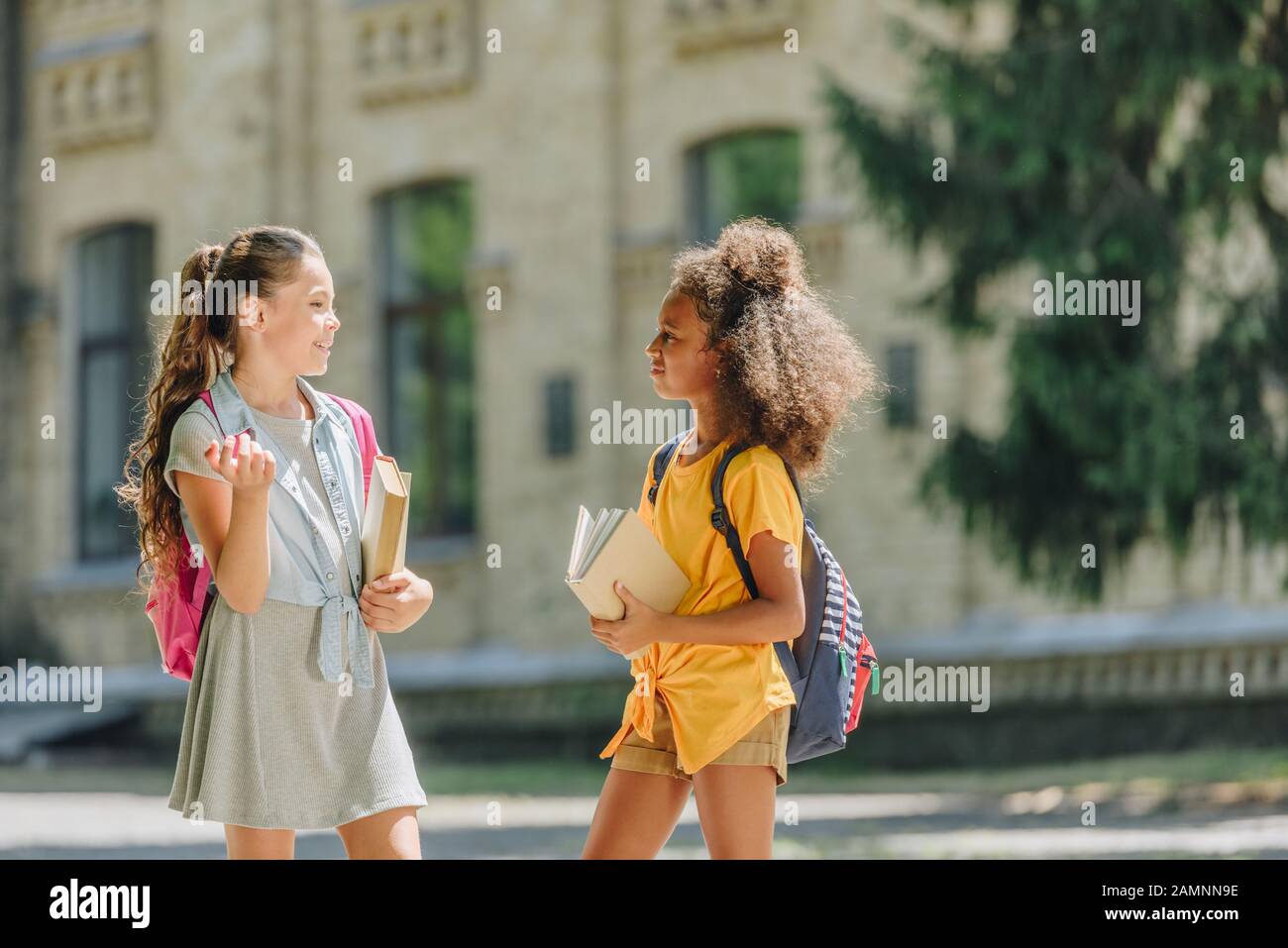 due allegre studentesse multiculturali che tengono libri e parlano in cortile Foto Stock