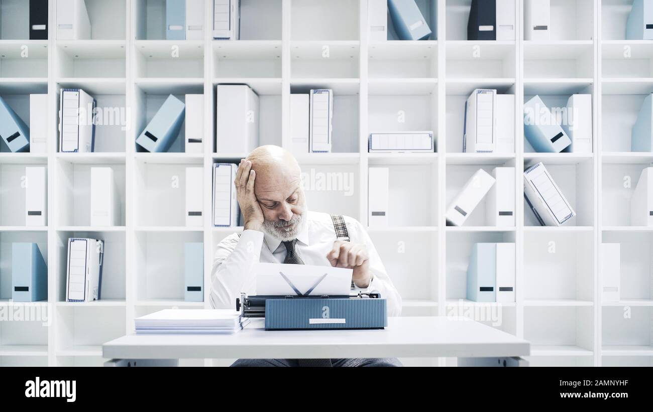 Uomo d'affari pigro senior o giornalista che lavora in ufficio, è seduto alla scrivania e utilizzando una macchina da scrivere Foto Stock
