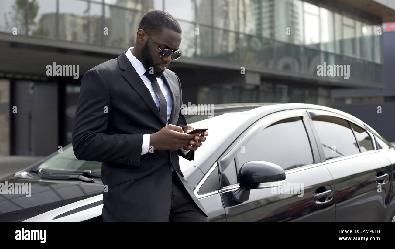 Uomo sicuro che digita e-mail sullo smartphone, in piedi vicino alla sua auto di lusso Foto Stock